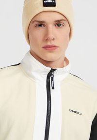 Crème en witte ritsvoering fleece jas met zwarte accenten. Beigebruine gebreide beanie met een zwart label, gladde textuur en een aansluitend ontwerp.