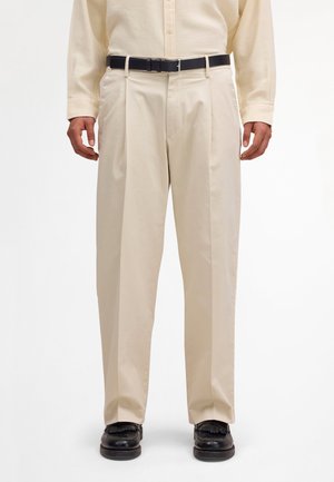 Beige plissierte Hose, getragen mit einem schwarzen Gürtel, beigem, geknöpftem Hemd und schwarzen Leder-Loafers bei einer stehenden Person.