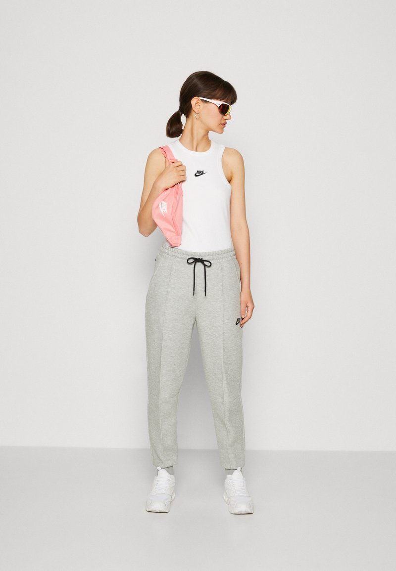 Camiseta de tirantes Nike en blanco con logo negro, combinada con pantalones de chándal gris claro con cordón. El modelo sostiene un bolso rosa y lleva gafas de sol.