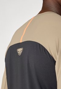 Rövid ujjú sport póló bézs és fekete szövetből, egyszínű hátsó résszel, narancssárga díszcsíkkal és logóval a háton.