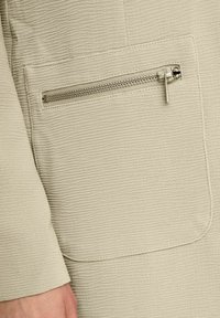 Beżowa teksturowana tkanina z poziomym wzorem prążków i detalem kieszeni na zamek. Kieszeń posiada metalowy zamek z suwakiem.