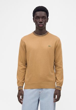 Mann mit kurzen lockigen Haaren, der einen beigefarbenen Langarmpullover mit schwarz-beige gestreiften Bündchen und Kragen trägt, kombiniert mit hellblauen Jeans.