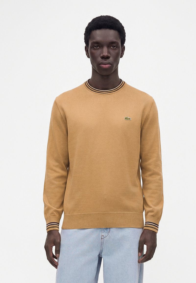 Homme aux cheveux courts et bouclés, portant un pull à manches longues beige avec des poignets et un col rayés noirs et beiges, associé à un jean bleu clair.
