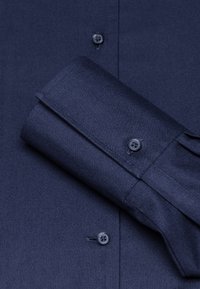 Camicia in cotone blu navy con chiusura a bottoni, polsino con un solo bottone e una texture liscia. Manica piegata mostrata in evidenza.