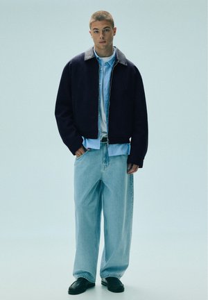 Jeune homme debout portant une veste noire sur une chemise bleu clair, un jean large bleu clair et des chaussures noires sur un fond uni clair.