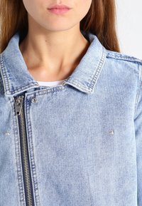 Světle modrá džínsová bunda se zipem, špičatým límečkem a stříbrnými kovovými detaily. Zničená textura, detailní pohled.