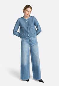 Giacca blu trapuntata con scollatura rotonda, due tasche frontali e bottoni neri. Abbinata a jeans in denim chiaro a gamba larga e scarpe nere a punta.
