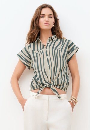 Pitkäaikainen nainen, jolla on aaltoilevat hiukset. Hänellä on päällään lyhythihainen, vihreä-beige raidallinen nappipaita, joka on solmittu vyötärölle, ja valkoiset korkeavyötäröiset housut.