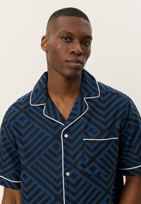 Camisa de manga curta em padrão geométrico preto e azul, apresentando um bolso no peito e um detalhe em branco ao longo do colarinho e da pala dos botões.