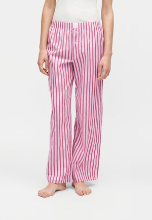 Személy laza, piros-fehér, függőleges csíkos pizsamanadrágot visel, rugalmas derékrésszel és gombos sliccel, mezítláb áll egyszínű háttér előtt.