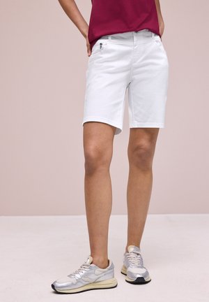 Person iført hvide knælange shorts, bordeaux top og sølv sneakers står på lyse gulv mod lys baggrund.