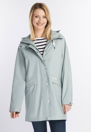 Ung kvinde smilende iført en lyseblå regnjakke over en sort-hvid stribet skjorte med mørke jeans mod en ensfarvet hvid baggrund.