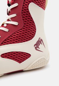 Αθλητικό παπούτσι ψηλού τύπου με κόκκινα διχτυωτά πάνελ, μπεζ επικάλυψη, λευκά κορδόνια και μπορντό λογότυπο σε σχήμα δοντιού στη φτέρνα και το μοτίβο της σόλας.