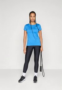 Camiseta deportiva azul de manga corta, combinada con mallas negras. La modelo lleva zapatillas negras y sostiene una cuerda para saltar, con un fondo contrastante.