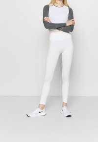 Leggings blancos ajustados y una camiseta blanca de manga larga con detalles en gris. La camiseta presenta detalles de malla en las mangas. Se lleva con zapatillas blancas.