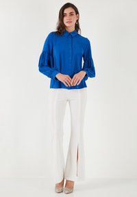 Blusa blu a maniche lunghe con bottoni frontali e maniche a palloncino elastiche; abbinata a pantaloni bianchi a vita alta con fessure laterali.