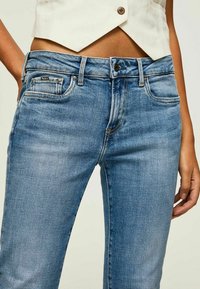 Jeans de mezclilla azul claro con un diseño ajustado, estilo de cinco bolsillos y costuras visibles. La tela tiene una textura suave con un leve desgaste.