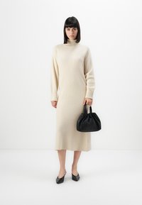 Una donna che indossa un lungo vestito a pullover beige con collo alto, tiene una borsa nera arricciata e indossa scarpe nere con tacco basso su uno sfondo bianco.
