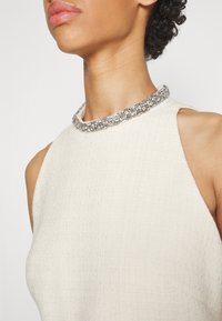 Femme portant un haut sans manches écru texturé avec un collier ras du cou étroit et étincelant en strass.