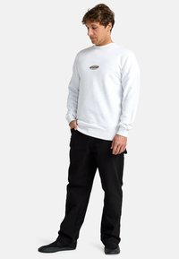 White sweatshirt with round logo in center, long sleeves, ribbed cuffs and hem, paired with black pants and black footwear.