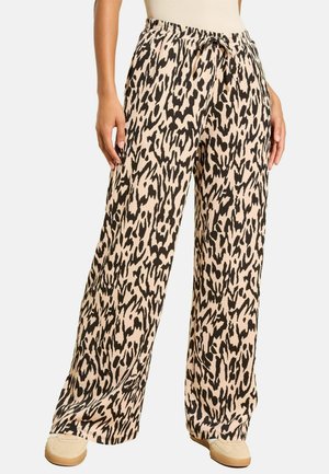 Pants ar plašām kājām, melnbaltu abstraktu leoparda rakstu, ar mežģījuma jostu, valkā vienādu beigu sporta apavu un baltu bezdarbības topiņu.