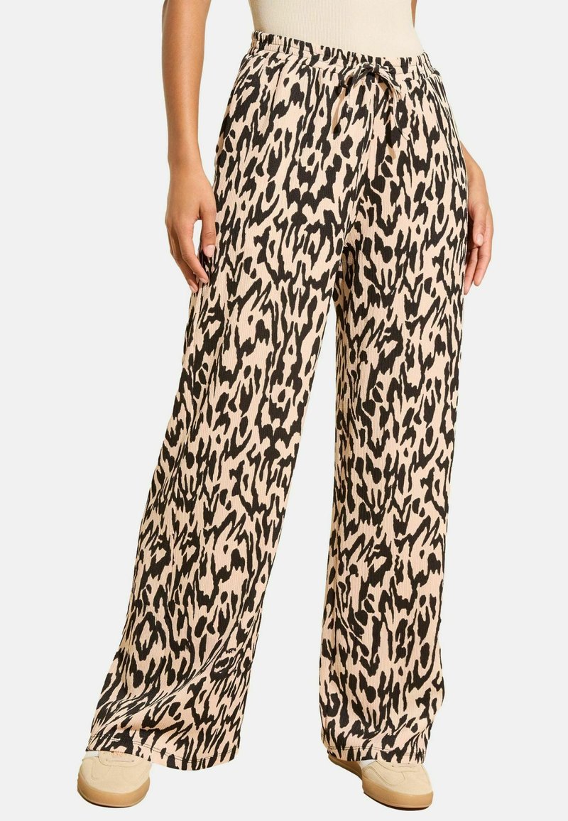 Pants ar plašām kājām, melnbaltu abstraktu leoparda rakstu, ar mežģījuma jostu, valkā vienādu beigu sporta apavu un baltu bezdarbības topiņu.