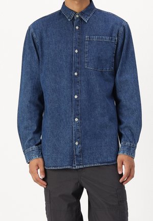 Mężczyzna ubrany w niebieską, długorękawową koszulę jeansową zapinaną na guziki z kieszenią na piersi oraz ciemne spodnie typu cargo, stojący na białym tle.