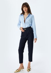 Camisa de botonadura azul claro con cuello, combinada con pantalones de navy y tacones de punta dorados tipo slingback. Presenta un cinturón negro con hebilla dorada.