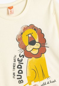 Cremefarbenes Baumwoll-T-Shirt mit einem Cartoon-Löwen mit einer braunen Mähne, begleitet von dem Text "Spaß mit Freunden" darunter.