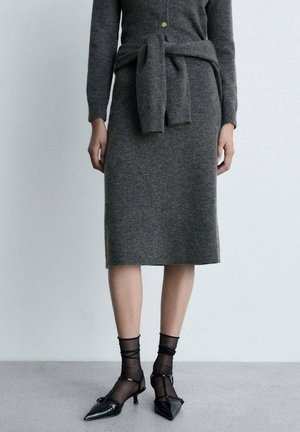 Femme portant une jupe en maille grise avec un pull assorti noué à la taille, des chaussettes transparentes noires jusqu'à la cheville et des chaussures noires à talons bas et bouts pointus.