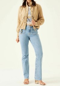 Béžová zkrácená bunda s elastickými manžetami, světle modré džíny s vysokým pasem a zlaté sandály na blokovém podpatku na světlém pozadí.