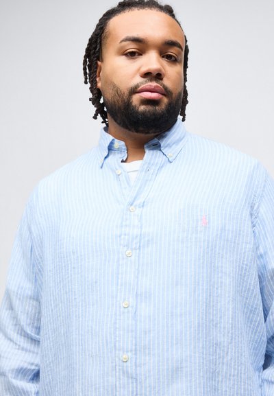 Άνδρας με γενειάδα και dreadlocks, φορώντας μπλε και λευκό ριγέ πουκάμισο πάνω από λευκό τοπ, κοιτάζοντας μπροστά με ουδέτερη έκφραση.