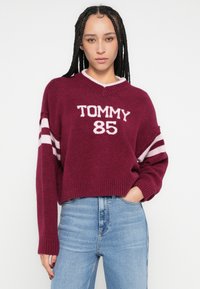 Burgundia värvi kootud kampsun V-kaelusega, millel on tekst "TOMMY 85", roosade ja valgete triibuliste varrukatega ning soonikkoes äärega. Kombineeritud siniste teksadega.