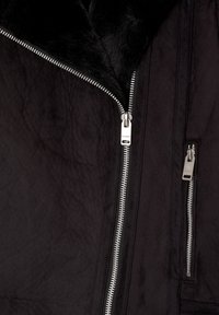 Chaqueta negra con cremallera que presenta un cuello de piel sintética, tejido texturizado, herrajes plateados y múltiples bolsillos con cremallera en la parte delantera.
