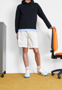 Svart huvtröja, ljusblå skjorta under, vita shorts och vita sneakers med blå detaljer. Står bredvid en stol med orange detaljer.