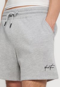 Shorts décontractés gris clair avec taille à cordon et poches latérales, portés par une personne avec une main dans la poche, comportant un petit logo noir sur la jambe.