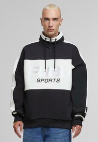 Sort og hvid pullover sweatshirt med høj krave, halv lynlås, stort "FUBU SPORTS" logo og ribbede ærmer. Glat stofstruktur.