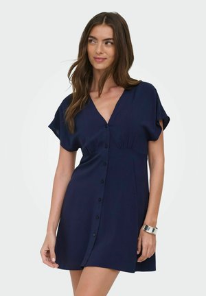 Femme portant une robe courte bleu marine boutonnée sur le devant, à manches courtes, avec un bracelet manchette en argent, regardant de côté avec un léger sourire.