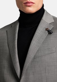 Mann, der ein graues kariertes Sakko über einem schwarzen Rollkragenpullover trägt, mit einem kleinen braunen Knopf und einem schwarzen Stoffetikett am Revers des Sakkos.