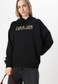 Črna kapucar iz mehkega blaga z osrednjim logotipom "CALVIN KLEIN" v svetli barvi. Ohlapen kroj z rebrastimi manšeti.