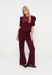 Vrouw in een bordeauxrode pailletten top met pofmouwen en bijpassende wijde broek, staand met één hand op de heup tegen een witte achtergrond.