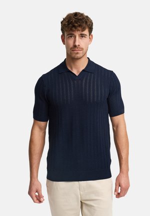 Mann mit lockigem Haar, der ein marineblaues kurzärmeliges Strick-Poloshirt und beige Hosen vor einem schlichten Hintergrund trägt.