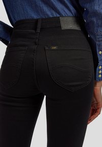 Gros plan d'une personne portant un jean noir Lee et une chemise en denim bleu avec des boutons visibles sur la manche.