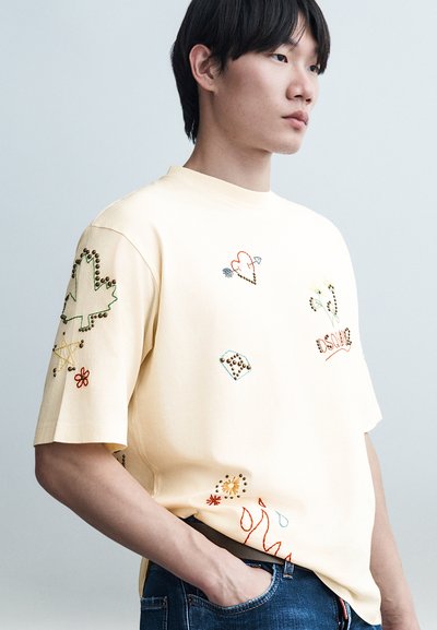 Jeune homme portant un t-shirt crème oversize avec des motifs colorés brodés et cloutés, une main dans la poche de son jean bleu.