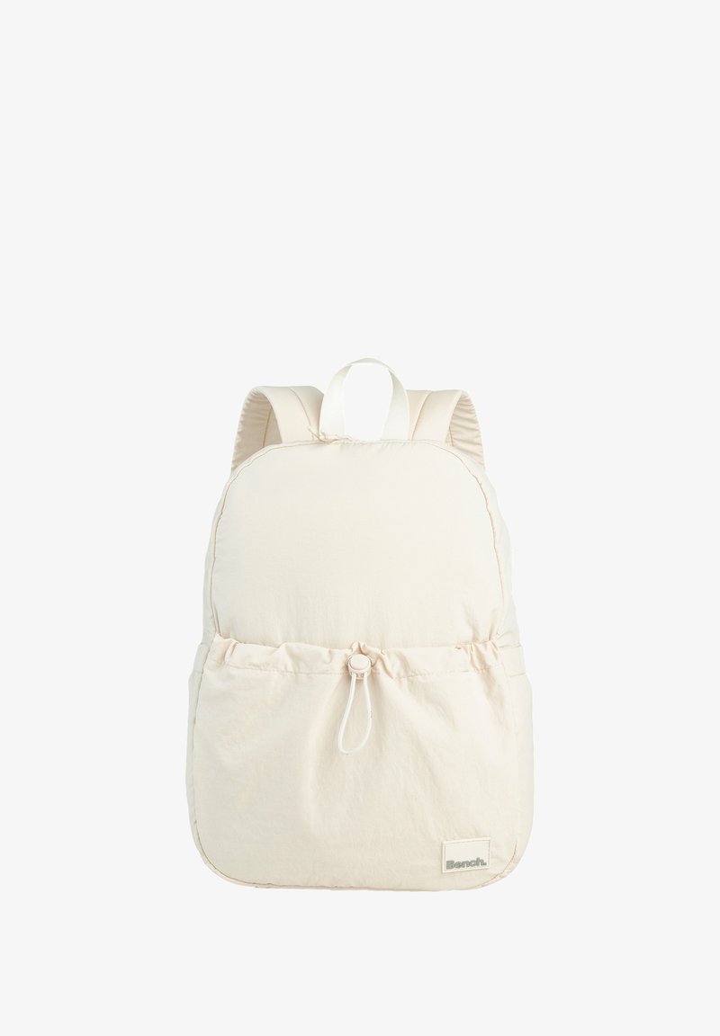 Cream-colored backpack with a rounded shape, soft fabric, adjustable straps, and a front pocket with a drawstring closure. Branding label visible.