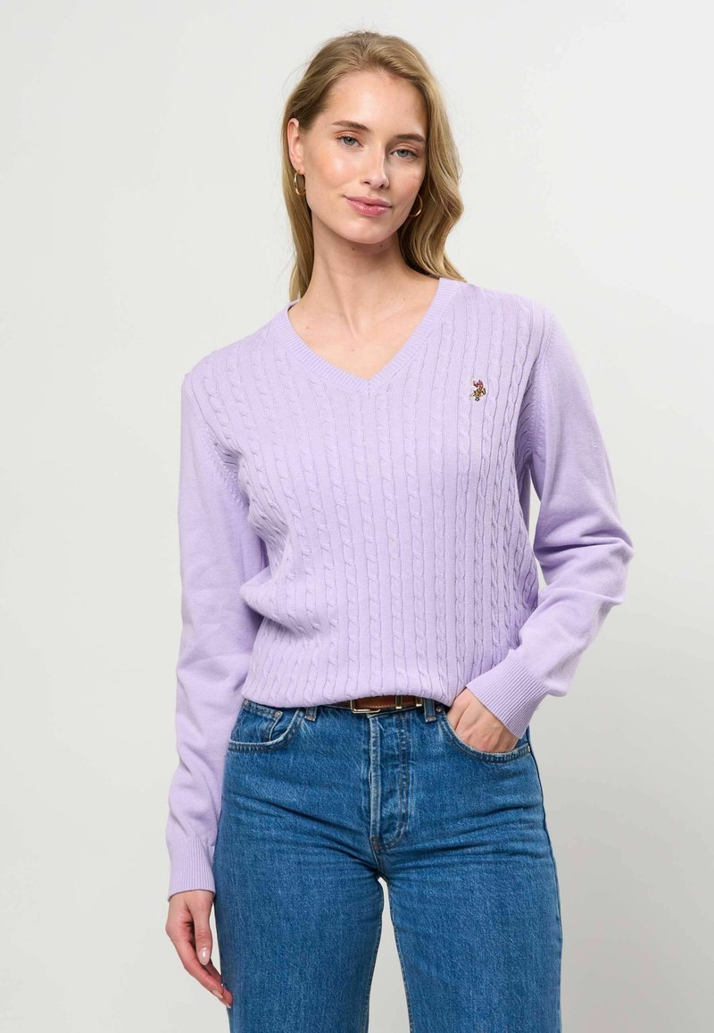 Femme aux cheveux blonds portant un pull en maille torsadée violet clair et un jean bleu, debout avec une main dans la poche sur un fond uni.