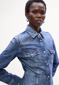 Camisa de denims com colarinho pontudo, dois bolsos no peito e acessórios prateados. Apresenta um tom azul claro e um tecido robusto e texturizado.
