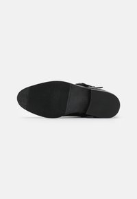 Zapato de cuero negro con suela plana, fondo de goma texturizada y un diseño minimalista. Cuenta con una hebilla ajustable para el cierre.