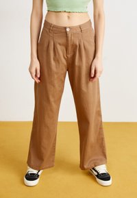 Brune vidde bukser med frontfolder, knappestenging og sidelommer, paret med svarte og hvite sneakers på en gul gulv.