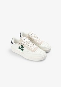 Zapatillas bajas blancas con acentos de ante beige, bordado de calavera y tibias verdes en los lados, cordones blancos y lengüetas en azul marino.